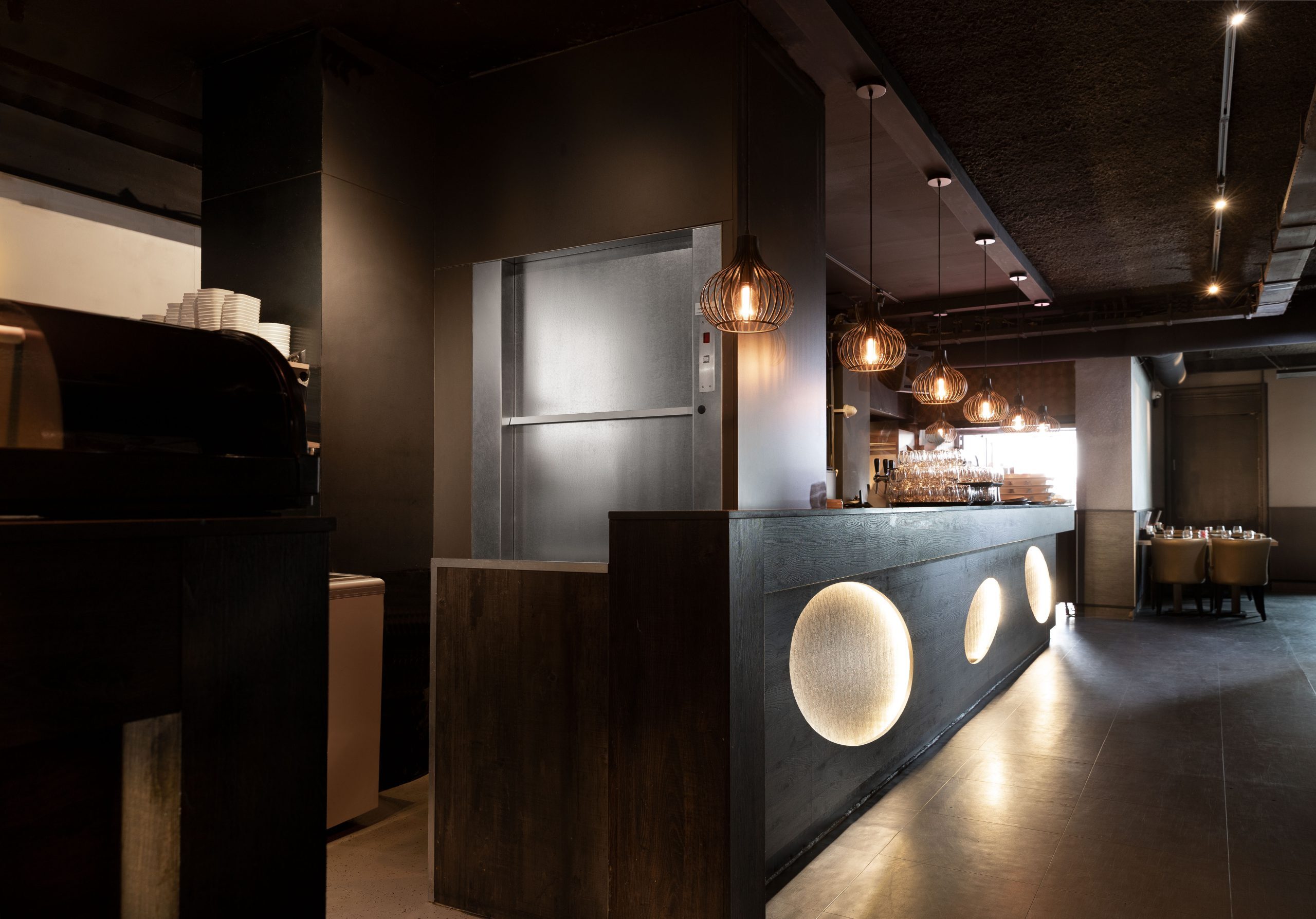 Moderne bar met houten elementen en sfeervolle verlichting. Achtergrond toont een lift.